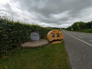 Welcome to Durrow sign in stonework along side the N77 road. Beside it is a round hay bale decorated for the scarecrow festival.