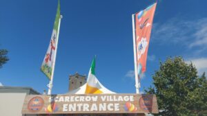 The entrance to the Durrow Scarecrow Festival is flanked by two tall scarecrow flags with a blue sky backdrop suggesting a fair weather day is being enjoyed by all in attendance.